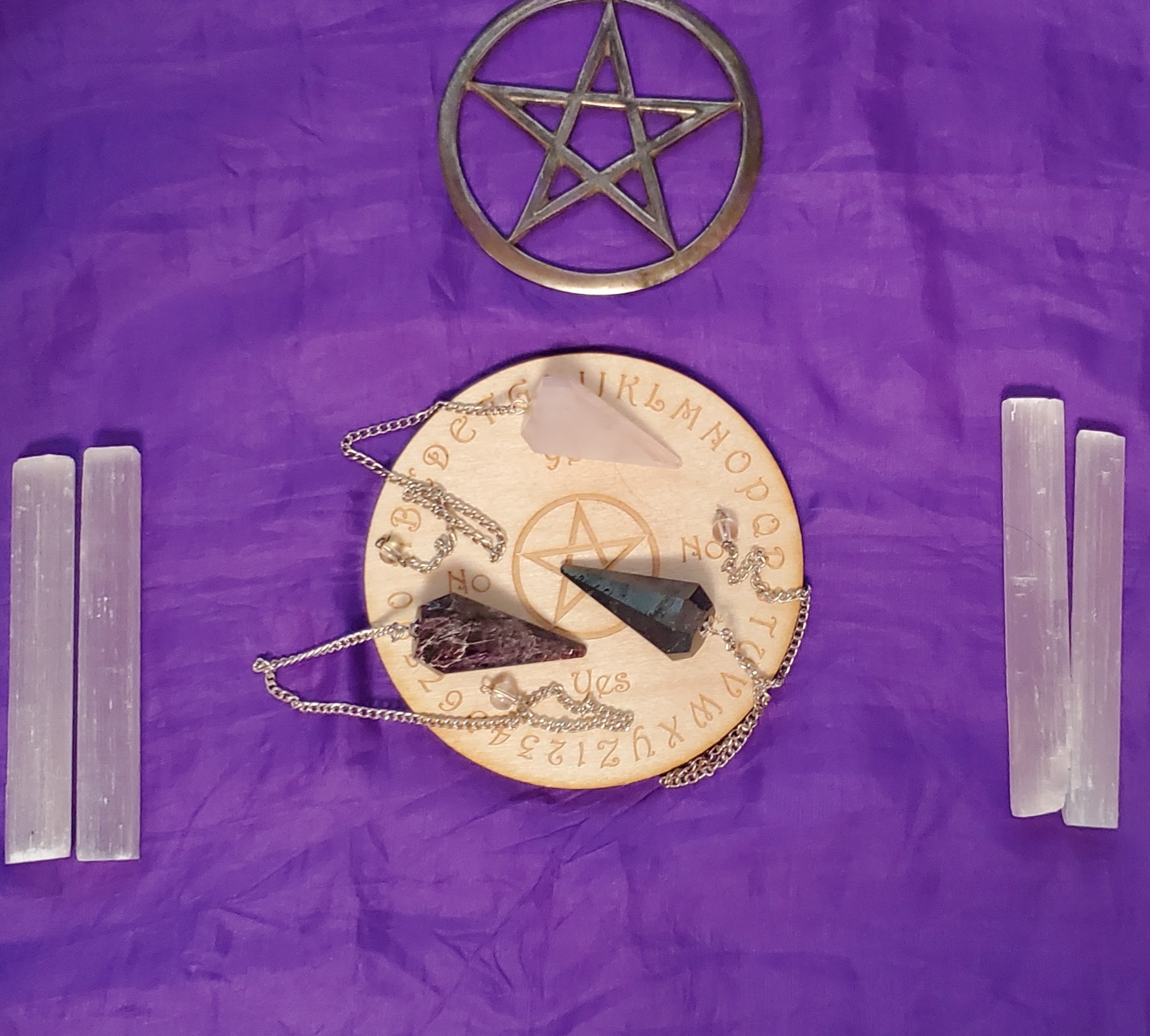 Silver pentacle center top above a wooden pendulum board with three pendulums on it, garnet, hemetite, and rose quartz. On each side are two selenite sticks
