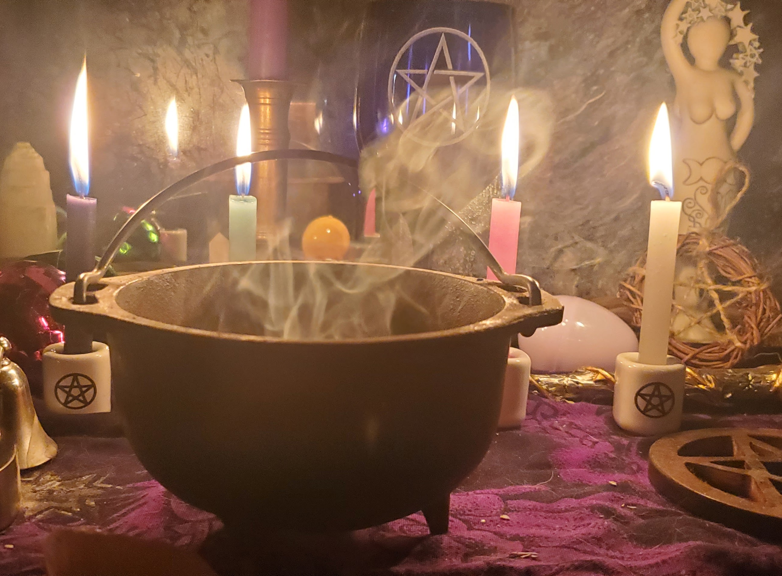 Di's cauldron smoking with candles lit behind it, a goddess statue to the back right, a wooden pentacle center right