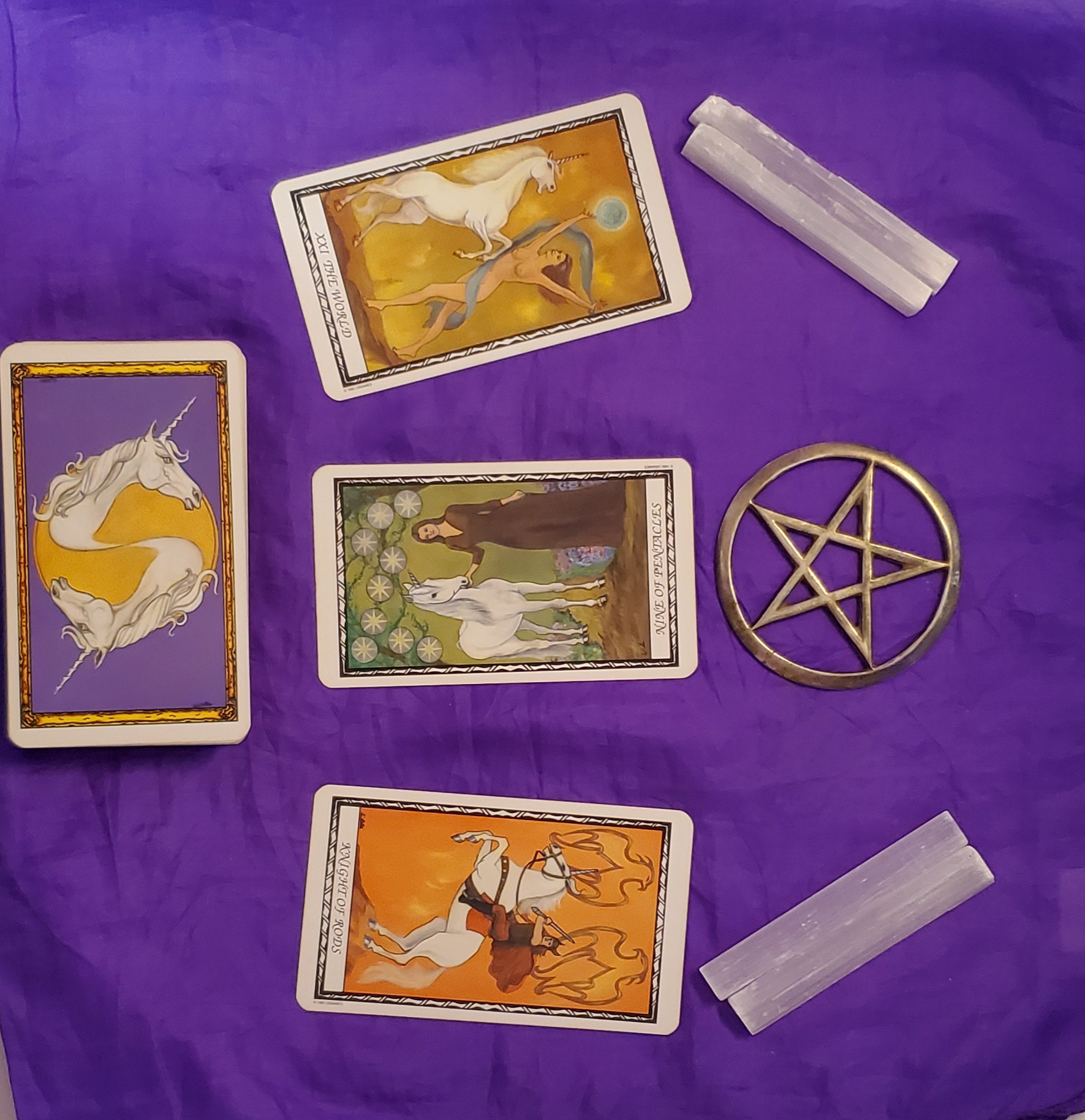 Right side center is a deck of unicorn tarot cards, face down. 3 tarot cards are drawn next to this. On the right is a silver pentacle, with 4 selenite sticks