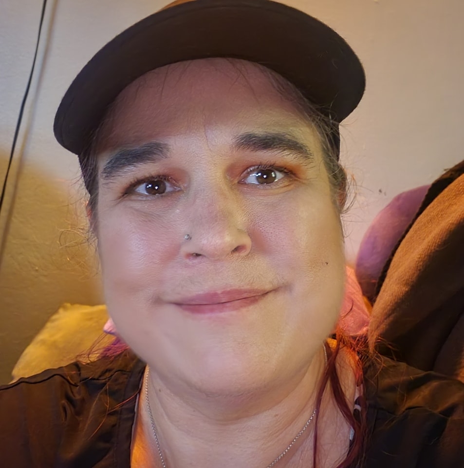 Image of Di the Yoga Witch smiling at the camera. Di is a middle aged woman with dark eyes, thick eyebrows, and pierced right nostril.
