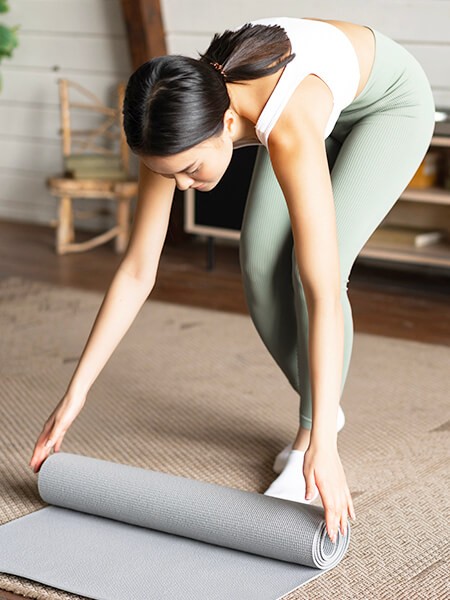 A woman with dark hair in a white sport bra and sage green yoga pants laying out a gray yoga mat on beige carpet
